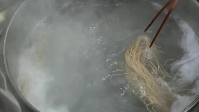 A close-up shot capturing the process of boiling noodles or spaghetti in a metal pot. The boiling water is bubbling vigorously with steam rising as the noodles are being cooked to an al dente texture 