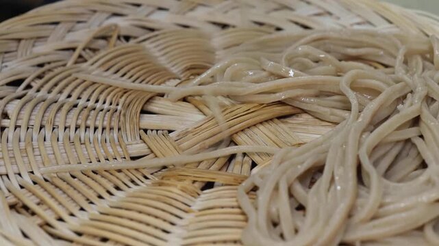 Close-up footage of raw, artisanal Japanese soba noodles (buckwheat noodles) carefully arranged on a traditional round woven bamboo tray (zaru). This high-quality shot highlights the delicate texture 
