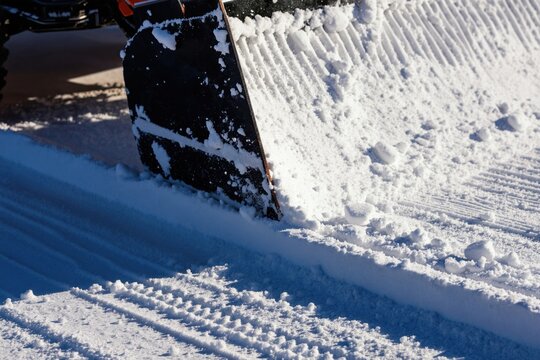 Close-up of snowplow blade clearing deep snow creating sharp grooves. Sunlight reflects on cleared path. Machine moves snow on a bright winter day. Road maintenance in action.