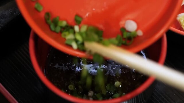 Close-up slow-motion footage of sliced green onions being added from an orange bowl into a dark, savory Asian soy-based dipping sauce. This versatile condiment is commonly used for dumplings, potstick