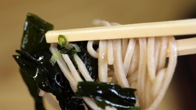 A macro shot focusing on traditional Japanese buckwheat soba noodles being lifted by bamboo chopsticks. The noodles are accompanied by dark green, glistening wakame seaweed and a garnish of finely sli