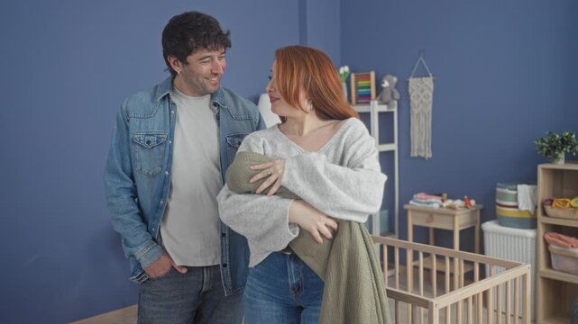 Couple cradling a swaddled baby in a home nursery with wooden crib, mobile and shelves; new parent joy.
