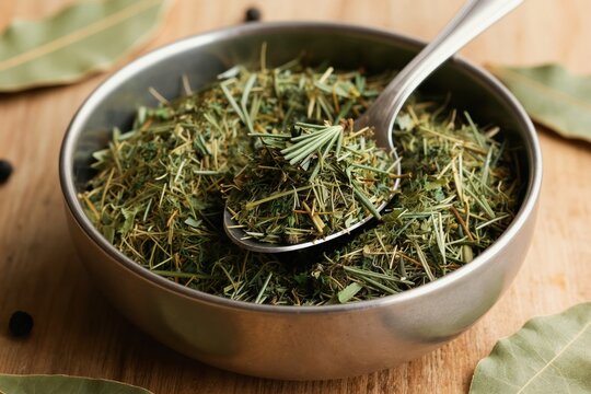 Dried culinary herbs, including rosemary and cilantro, in metal bowl with spoon. Bay leaves and peppercorns nearby on wooden surface. Ingredients for cooking and healthy meals.