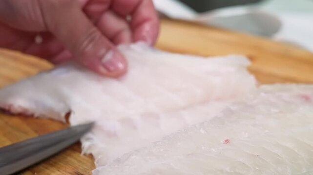 High-angle, close-up shot of a professional chef's hands carefully using a knife to fillet a fresh piece of white fish (resembling sea bass or cod) on a wooden cutting board. The video captures the pr