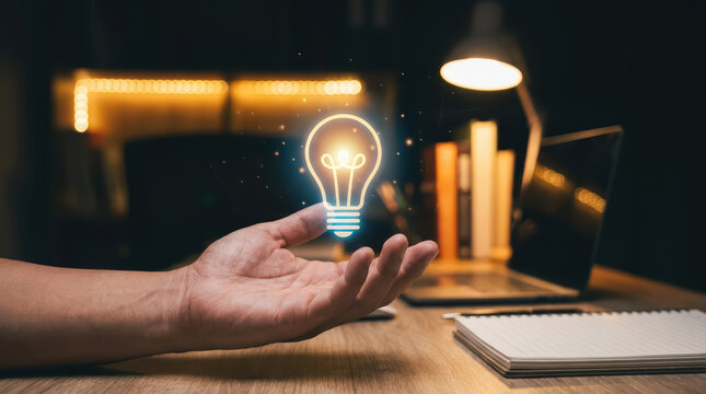 Hand holding glowing light bulb hologram, creative idea concept with laptop and notebook on wooden desk in dark room
