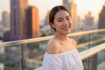 Obraz premium Portrait of a happy beautiful asian woman smiling on a rooftop with a city view at sunset