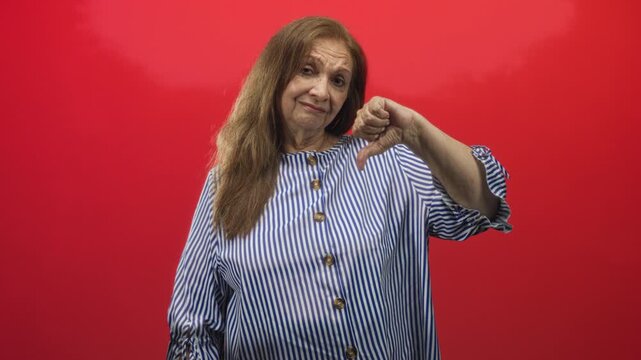 Woman senior hispanic showing thumb down gesture with visible thumb wearing blue striped button blouse in studio; disapproval.