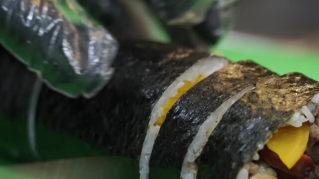 High-angle close-up of a chef slicing a traditional Korean kimbap roll into bite-sized pieces. The roll is filled with seasoned beef, yellow pickled radish, and fresh vegetables wrapped in white rice 