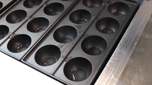 Close-up footage of a chef carefully oiling a traditional 28-hole iron takoyaki pan. The video captures the preparation process of making authentic Japanese octopus balls in a commercial kitchen setti