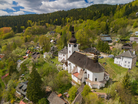 Aerial view of the historic Church of the Conversion of the Lord and traditional houses surrounded by lush green forests and rolling hills in Spania Dolina, Banska Bystrica Region, Slovakia.