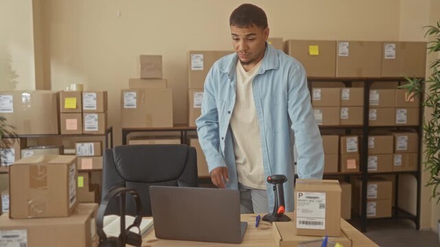 Man touching neck beside stacked shipping boxes, laptop and barcode scanner at a table in studio; concern entrepreneurship.