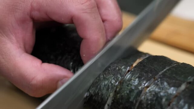 Close-up of a skilled chef&rsquo;s hand using a sharp Japanese kitchen knife to precisely cut a sushi roll wrapped in nori seaweed. This footage highlights the culinary artistry, fresh ingredients, and prof