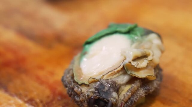 A high-quality close-up shot of a fresh raw abalone in its natural shell, being held and prepared. This premium seafood delicacy, highly valued in Asian cuisines like Japanese sashimi and Chinese fest