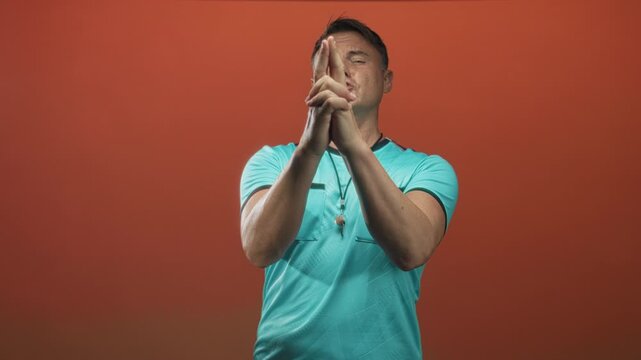 Man referee with whistle presses hands together into a finger gun gesture and winks while wearing turquoise shirt in studio; playful confidence.