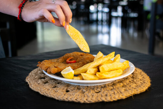Primer plano de una mano femenina tomando una papa frita de un plato de milanesa con papas en un entorno de restaurante, estilo de vida.