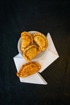 Vista cenital de empanadas fritas dispuestas sobre un plato blanco y servilletas, con un fuerte contraste sobre fondo de mesa negro r&uacute;stico.
