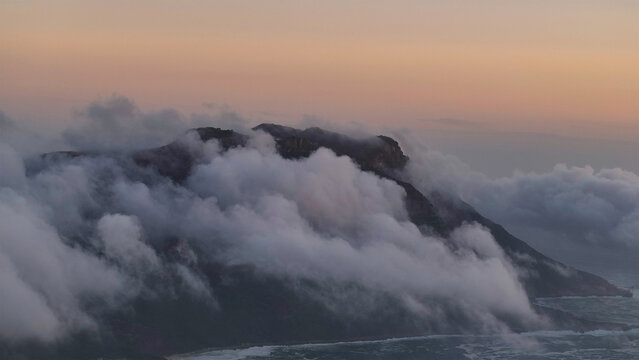 Aerial view of Table Mountain shrouded in thick white clouds rolling over the peaks during a soft sunset in Cape Town, Western Cape, South Africa.