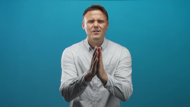 Man with hands pressed together in a pleading gesture facing camera in studio blue backdrop; pleading hope.