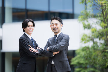 Fototapeta premium 屋外のビルの前で信頼感あるスーツの営業チームのポートレート法人営業と企業成長イメージカメラ目線の笑顔 上司と部下が腕を組む立つ