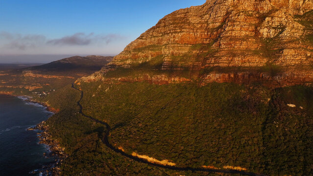 Aerial view of the winding Chapman's Peak Drive along steep rocky cliffs and lush green slopes during golden hour Cape Town, Western Cape, South Africa.