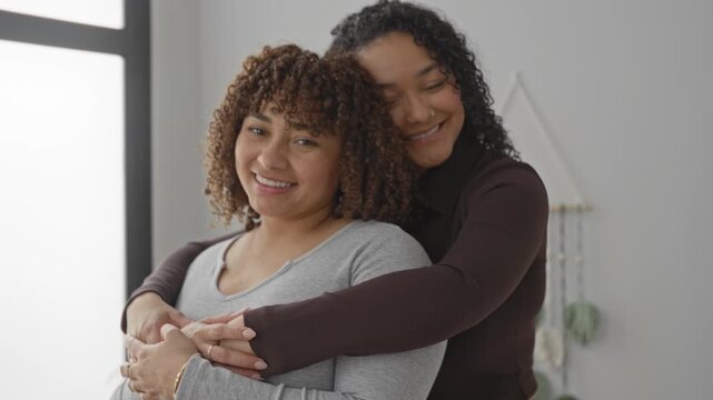 Two women smiling with hands clasped and arms hugging from behind by a window in an indoor building room; affection closeness.