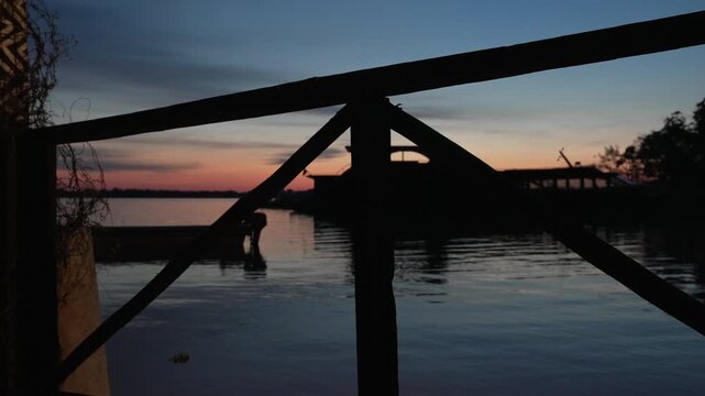 A silhouetted boat and structures are seen across the Amazon River at dusk, with vibrant sunset hues reflecting on the water. Ideal for travel or nature-themed projects.