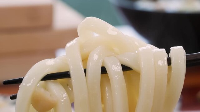 A detailed shot of steaming, thick white udon noodles being lifted by black chopsticks from a dark bowl. The noodles are served in a clear, savory dashi broth with garnishes, showcasing the chewy text