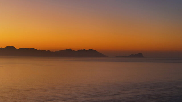 Aerial view of the silhouetted mountain range across the calm ocean waters under a vibrant orange and purple sunset sky in Cape Town, Western Cape, South Africa.
