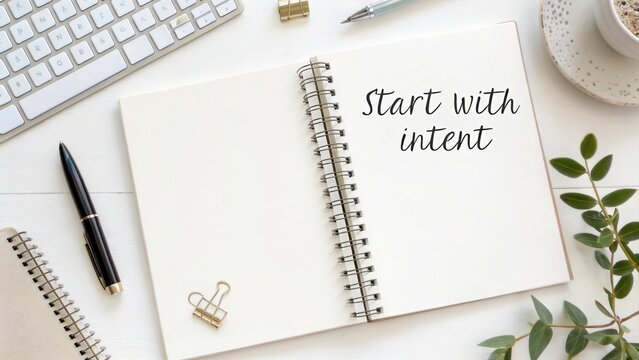 An open spiral notebook with the quote Start with intent written on its blank page, surrounded by office supplies.