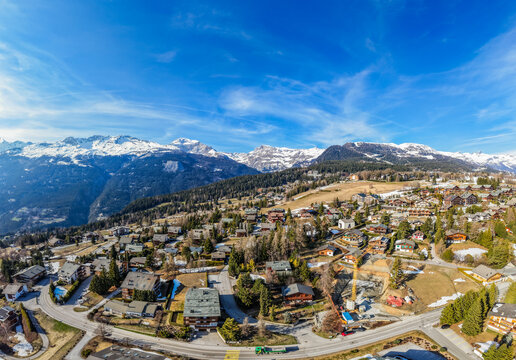 Aerial view of the alpine village with traditional chalets and pine trees against the backdrop of snow-capped peaks and a clear blue sky in Crans-Montana, Valais, Switzerland.