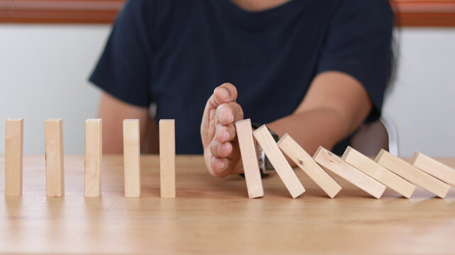 Close-up of hand stopping falling wooden domino blocks mid-action. Concept of risk management, crisis intervention, business continuity, and preventing chain reaction failure.