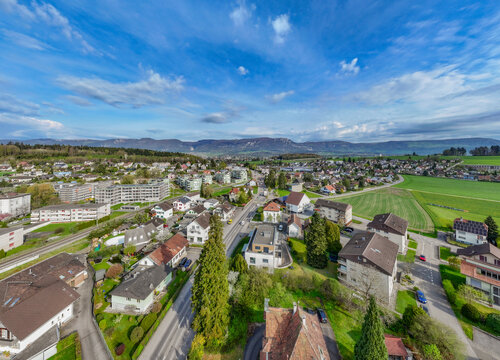 Aerial view of residential houses and apartment buildings surrounded by green fields and distant mountains under a blue sky in Biberist, Solothurn, Switzerland.