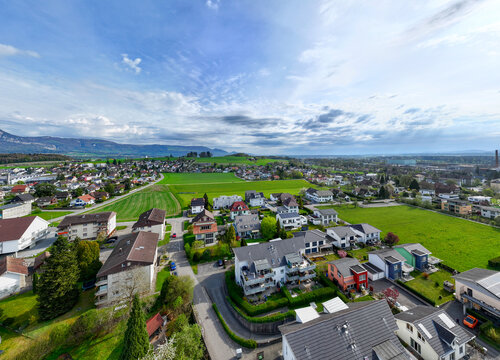 Aerial view of a residential neighborhood with modern houses, lush green fields, and distant mountains under a bright sky in Biberist, Solothurn, Switzerland.