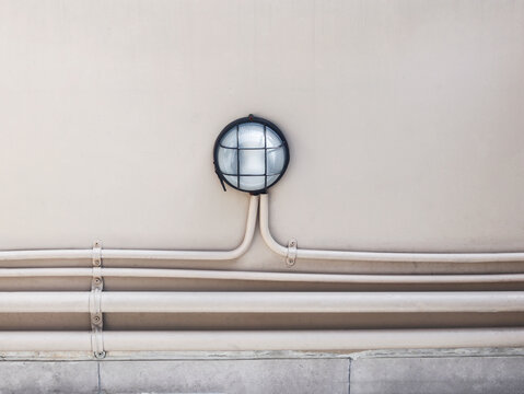 Industrial-style bulkhead wall lamp mounted on a beige exterior wall with visible horizontal conduit pipes. The round light features frosted glass enclosed in a protective metal cage.
