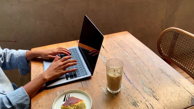 Close up of hands type on laptop keyboard with focused intent. Person wears light blue shirt, seated at wooden table.
