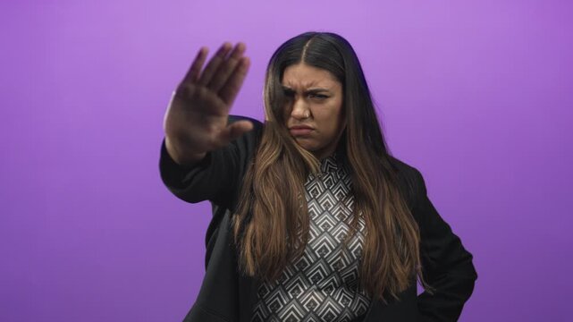 Woman hispanic plus size with raised hand showing palm out stop gesture in studio purple backdrop wearing patterned top and black jacket; defiance anger.