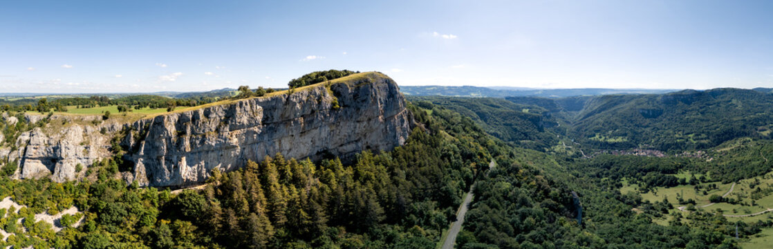 Aerial view of the massive limestone cliffs overlooking the lush Loue Valley with dense forests and a winding road under a clear blue sky in Mouthier-Haute-Pierre, Bourgogne-Franche-Comte, France.