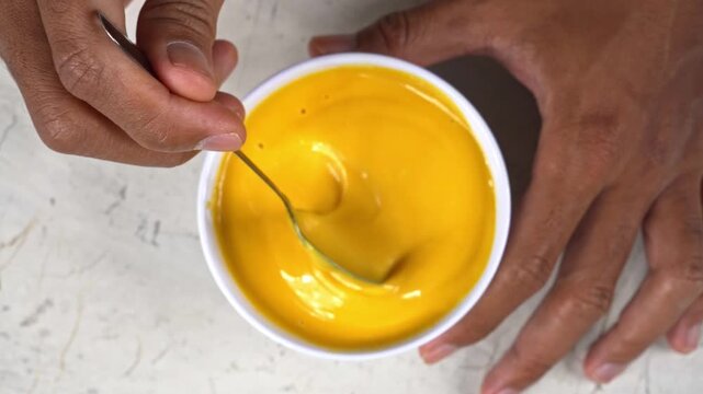 Top view of hand mixing smooth yellow mustard sauce in a clean white bowl on a light surface