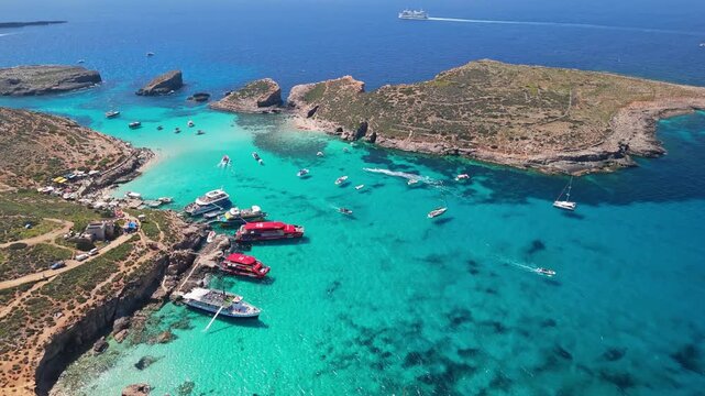 Blue Lagoon, Comino, Malta, features turquoise crystal-clear waters, rocky shores, and anchored boats near Cominotto island, viewed from above on a sunny day in the Mediterranean Sea.