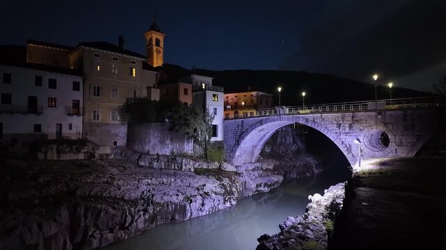 Kanal ob Soči, Slovenia, features its illuminated medieval stone arch bridge spanning the Soča River at dusk, with reflections on the water and historic riverside buildings in the old town.