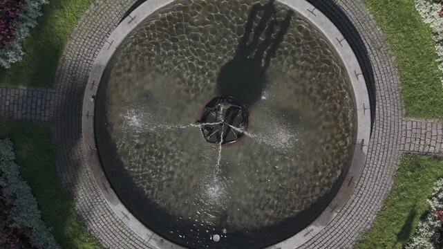 A captivating top-down drone shot of the Esplanadi Park Fountain in Helsinki, Finland, featuring a circular city fountain with crossing water jets, showcasing a peaceful and symmetrical landscape