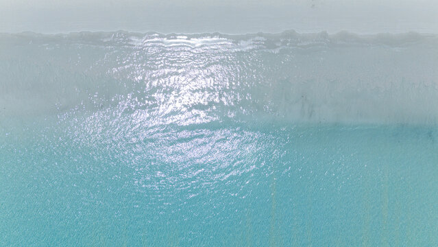 A serene aerial view of clear turquoise ocean water gently meeting a sandy beach, with sunlight reflecting on the waves.