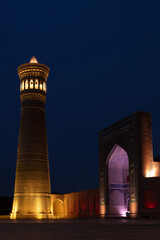 Obraz premium Kalyan Minaret and Kalyan Madrasah illuminated at night on Poi Kalyan Square in Bukhara. Dramatic golden and purple lighting highlights intricate brickwork and the pointed arch portal.