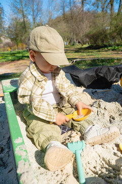 Toddler in beige cap and plaid shirt playing in sandbox at park on sunny spring day, blue sky and trees in background, outdoor childhood leisure.