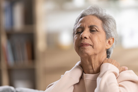 Senior woman feeling severe neck pain holding tight shoulder muscle suffering physical ache tension feeling stressed upset indoors resting alone on sofa having healthcare problem