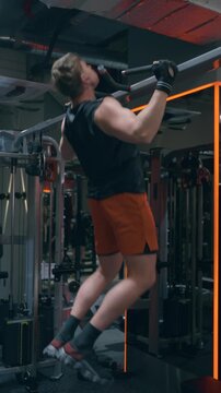 Muscular Caucasian man in his 30s performs wide-grip pull-ups targeting latissimus dorsi and upper back muscles on pull-up bar in modern gym - vertical, cinematic