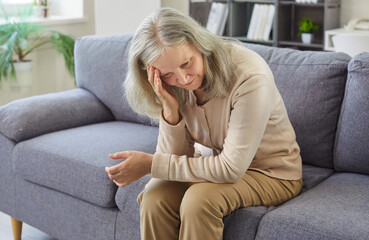 Obraz premium Sad elderly woman suffers from headache or migraine at home, sitting on sofa, facing health problem and pain, reflecting sick discomfort in a escape zone filled with senior care and distress.