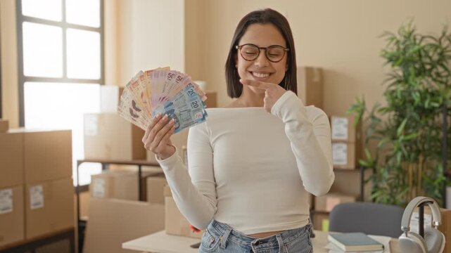 Woman holding colorful peso banknotes and delivery parcels amid stacked boxes in a building, smiles and points finger to cash; small business pride.