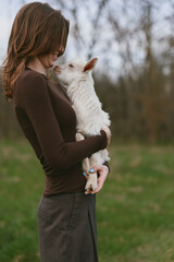 Naklejka premium Cheerful woman outdoors embracing a goat kid, warm moment of affection, natural light, rustic countryside background, intimate bond, calm expression, lifestyle portrait with gentle vibe