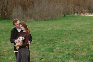 Naklejka premium A couple smiles in a sunlit park, one embracing a small dog. The scene captures warmth, companionship, and everyday romance in a bright, open green field.